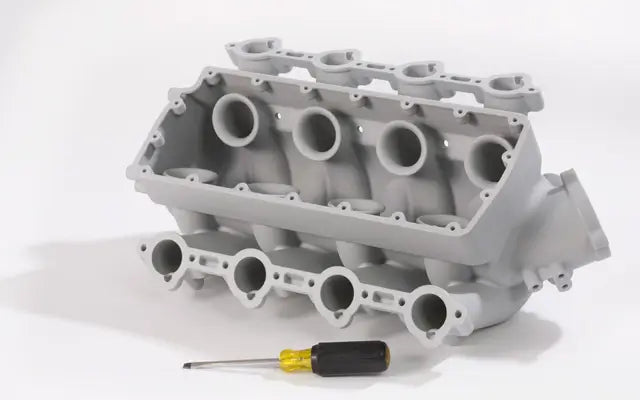 A 3D-printed automotive intake manifold held in hand with a miniature car and engine block nearby, showing rapid prototyping in the automotive industry.